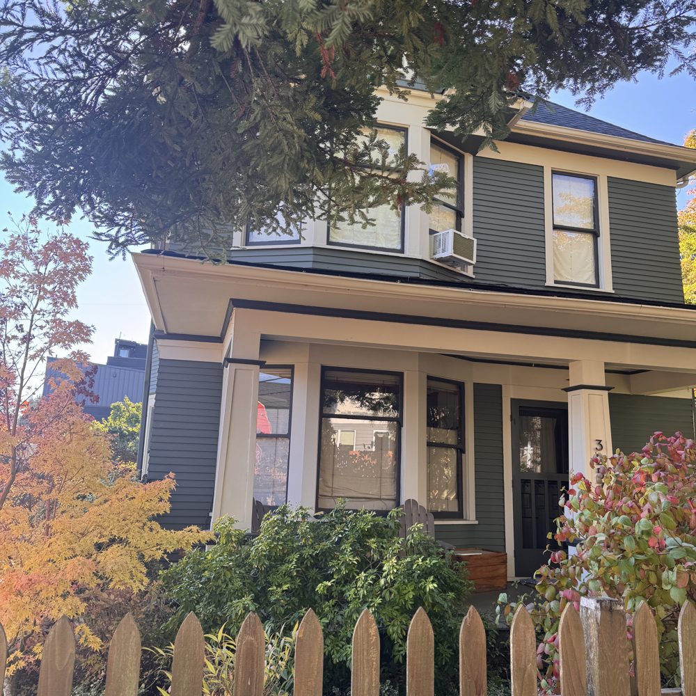 A gray early-1900s home with a deep porch behind a picket fence
