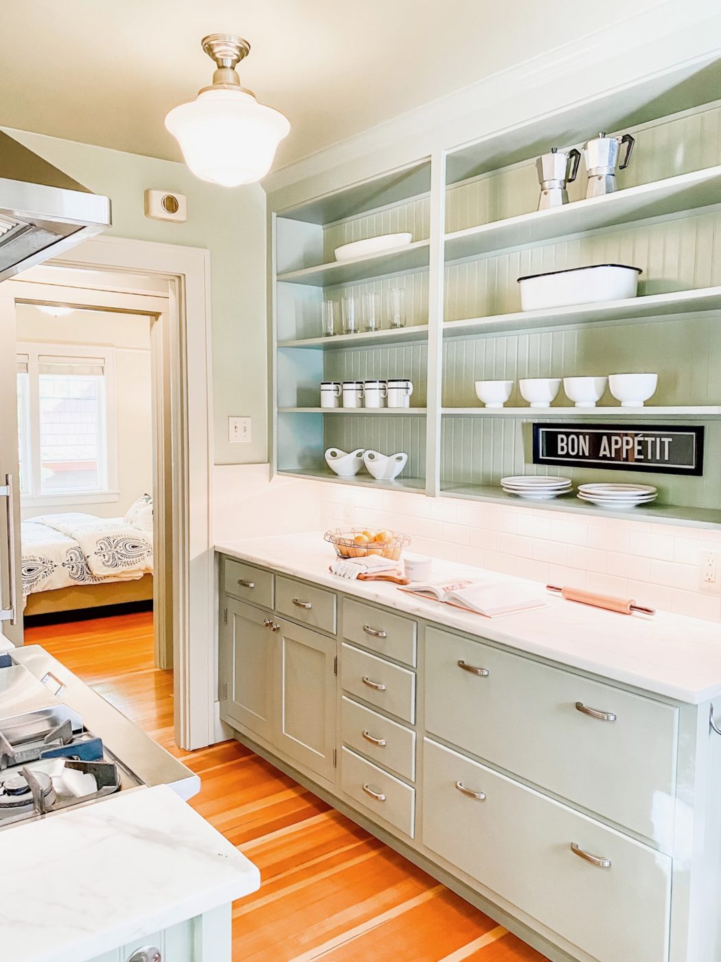 A sunny kitchen with sage cabinets and open shelves of white dishes