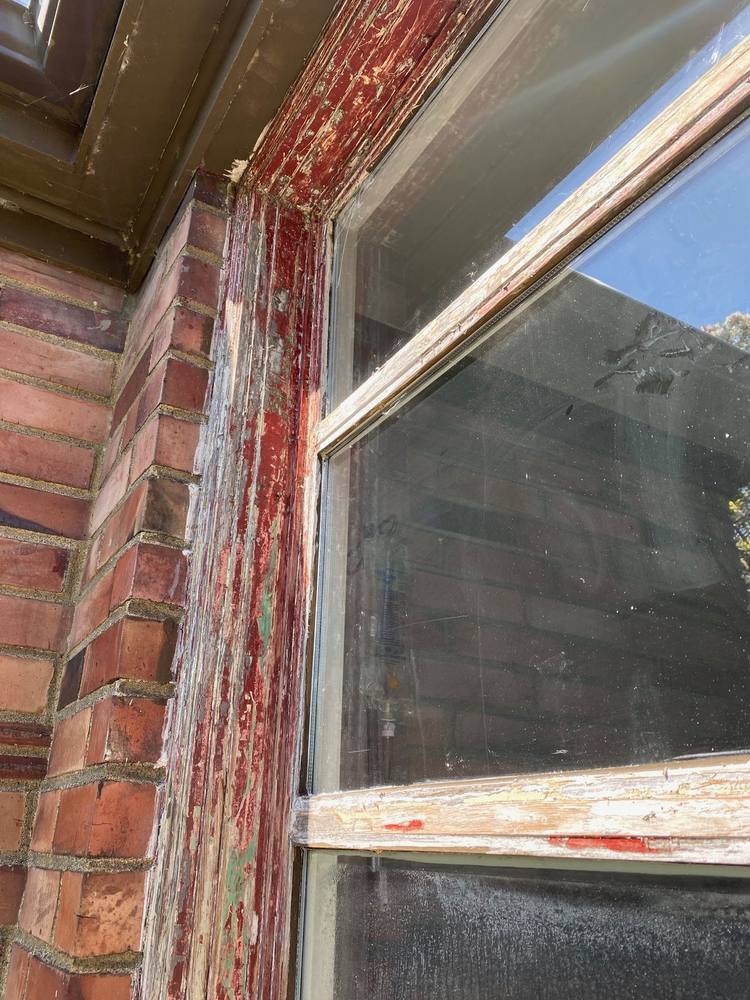 Layers of old red and green paint peeling from wood window trim on a brick home